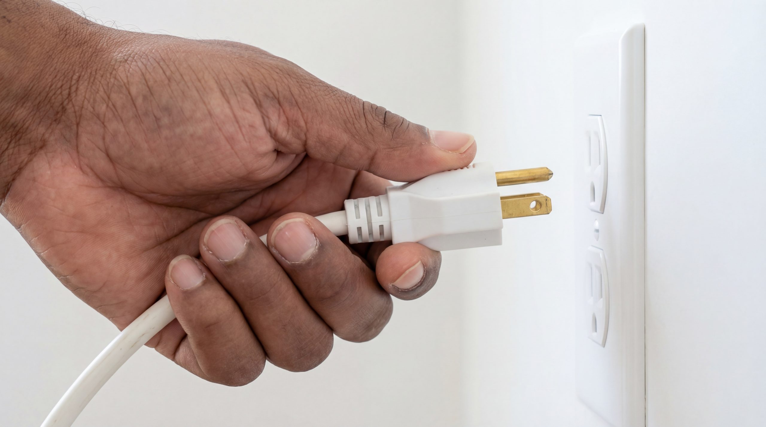 close up view of a black male hand inserting a white three prong electric plug into a wall outlet socket to connect power and energy photo